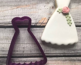 May include: A purple plastic cookie cutter shaped like a strapless wedding dress with a scalloped hem. Next to the cutter is a white iced cookie decorated with a pink flower and green leaves.