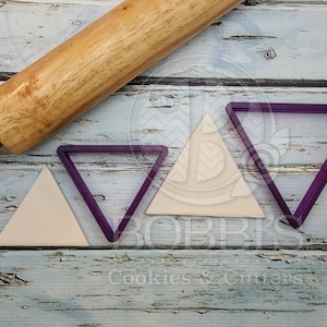 May include: Three purple triangle cookie cutters and a wooden rolling pin on a whitewashed wood surface.