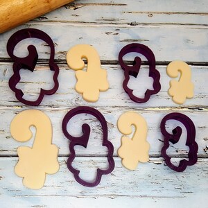 Puede incluir: Cortadores de galletas de pl&aacute;stico morado en forma de bastones de caramelo, con algunas galletas cortadas con los cortadores. Las galletas son de color beige claro.