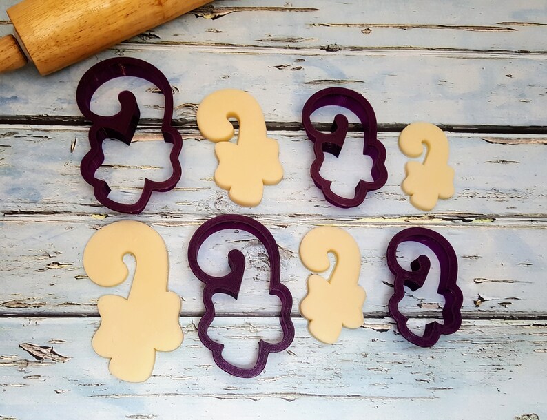 Puede incluir: Cortadores de galletas de pl&aacute;stico morado en forma de bastones de caramelo, con algunas galletas cortadas con los cortadores. Las galletas son de color beige claro.