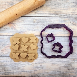 May include: A purple plastic cookie cutter set with a square frame, two heart shapes, and an X shape. The cookie cutter is on a wooden surface with a rolling pin and a piece of dough with the shapes cut out.