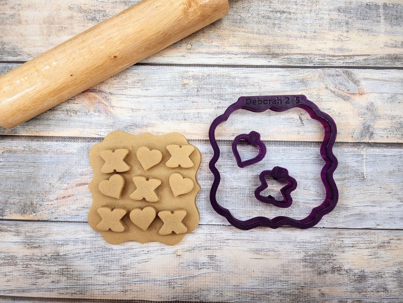 May include: A purple plastic cookie cutter set with a square frame, two heart shapes, and an X shape. The cookie cutter is on a wooden surface with a rolling pin and a piece of dough with the shapes cut out.
