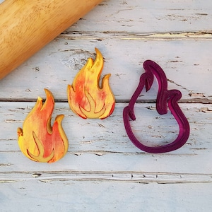 May include: Two yellow and orange flame-shaped cookies and a purple plastic cookie cutter in the shape of a flame. The cookies and cutter are on a white wooden surface.