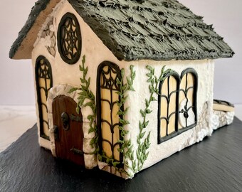 May include: A white gingerbread house with a green roof, decorated with ivy and black spiderweb windows. The house is sitting on a black slate surface.