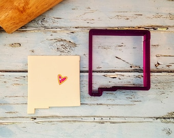 May include: A pink cookie cutter in the shape of the state of New Mexico. A small heart-shaped cookie is next to the cutter.
