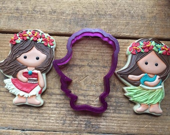 May include: Two decorated sugar cookies with brown hair and floral headbands, one wearing a red dress and the other a green grass skirt, with a purple plastic cookie cutter in the shape of a person.