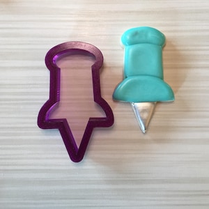 May include: A purple plastic cookie cutter and a turquoise blue sugar cookie in the shape of a push pin.