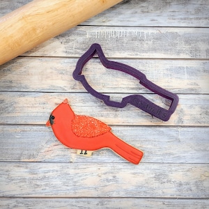 May include: A red cardinal cookie decorated with red sugar crystals, next to a purple plastic cookie cutter shaped like a cardinal with the text "Cardinal 5" on it.