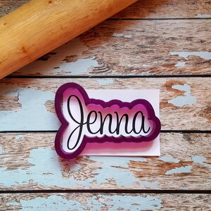 May include: Purple plastic cookie cutter in the shape of the name "Jenna" on a wooden surface.