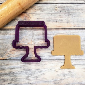 May include: Purple plastic cake stand cookie cutter with the text "Cake Stand 3 4" on the top. A brown sugar cookie in the shape of a cake stand is next to the cutter.