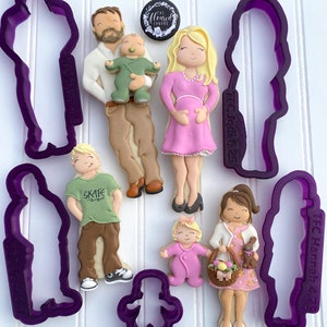 May include: Purple plastic cookie cutters in the shapes of a family: a man, a woman, a boy, a girl, a baby, and a toddler. The cutters are arranged on a white surface with decorated cookies in the same shapes.