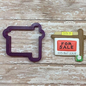 May include: A purple plastic cookie cutter shaped like a house, next to a decorated cookie shaped like a "For Sale" sign with a brown wooden frame, a red sign with white text, and a yellow "Sold" sticker.