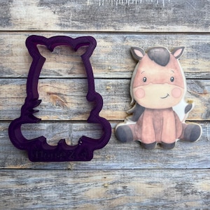 May include: Purple plastic cookie cutter shaped like a sitting horse. The cutter is next to a decorated sugar cookie with a brown horse design.