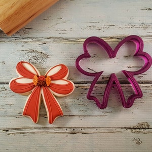 May include: A red and white bow cookie cutter and a decorated cookie in the shape of a bow. The cookie cutter is purple and the cookie is decorated with orange and white icing.