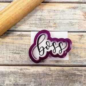 May include: A purple cookie cutter with the word "boss" in a cursive font. The cookie cutter is on a white piece of paper, with a wooden rolling pin in the background. The background is a light-coloured, weathered wooden surface.