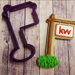 May include: Purple plastic cookie cutter shaped like a real estate sign with the letters KW on the sign. Next to the cutter is a decorated cookie with brown icing for the sign post and green icing for grass.