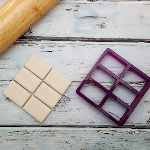 May include: A white fondant square divided into nine sections with a purple plastic mould for making chocolate bars on a white wooden surface.