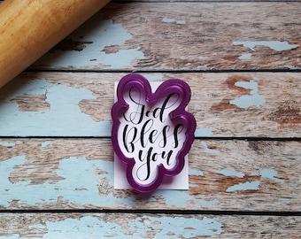 Könnte beinhalten: Lilafarbener Plätzchenausstecher aus Kunststoff mit den Worten "God Bless You" in einer kursiv geschriebenen Schrift.
