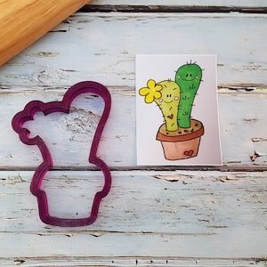 May include: A purple plastic cookie cutter shaped like a cactus plant in a pot. The cutter is on a white wooden surface next to a card with a drawing of a cactus plant.