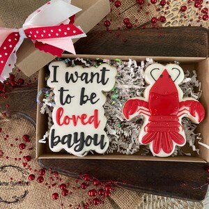K&ouml;nnte beinhalten: Eine Geschenkbox gef&uuml;llt mit wei&szlig;en und roten Keksen. Ein Keks ist mit dem Text "I want to be loved bayou" verziert und der andere Keks ist mit einem roten Krebs verziert. Die Box steht auf einer braunen Holzoberfl&auml;che.