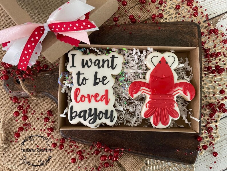 K&ouml;nnte beinhalten: Eine Geschenkbox gef&uuml;llt mit wei&szlig;en und roten Keksen. Ein Keks ist mit dem Text "I want to be loved bayou" verziert und der andere Keks ist mit einem roten Krebs verziert. Die Box steht auf einer braunen Holzoberfl&auml;che.