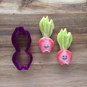 May include: Two pink and green decorated sugar cookies shaped like radishes with smiling faces. A purple plastic cookie cutter shaped like a radish is also shown.