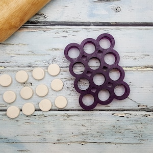 May include: A purple plastic cookie cutter shaped like a bunch of grapes with 18 individual circles.  There are 18 white circles of dough next to the cutter.
