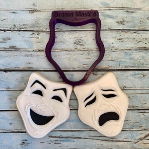 May include: Two white sugar cookies decorated with black icing to look like theater masks. One mask is smiling and the other is frowning. A purple plastic cookie cutter shaped like a theater mask is above the cookies. The cookie cutter has the text "Drama Mask'4" on it.