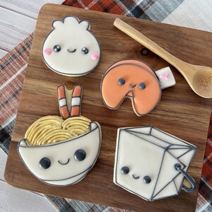 May include: Five decorated sugar cookies on a wooden board. The cookies are shaped like a bowl of noodles, a fortune cookie, a dumpling, a takeout box, and chopsticks. The cookies have cute cartoon faces and are decorated with icing.
