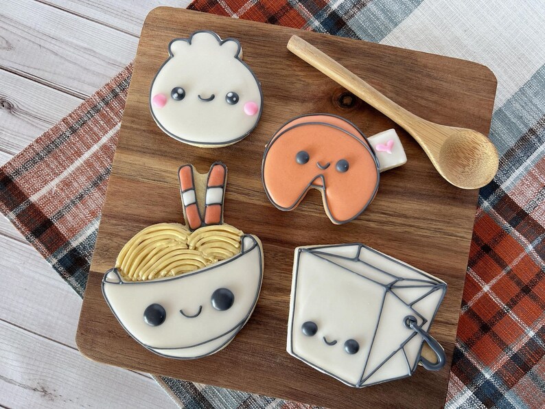May include: Five decorated sugar cookies on a wooden board. The cookies are shaped like a bowl of noodles, a fortune cookie, a dumpling, a takeout box, and chopsticks. The cookies have cute cartoon faces and are decorated with icing.
