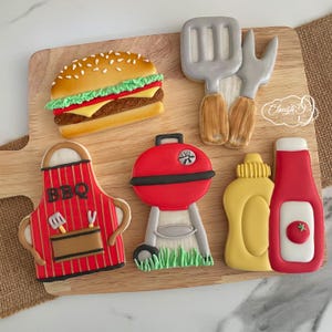 Pode incluir: Um conjunto de nove bolachas de açúcar decoradas com tema de churrasco. As bolachas incluem um hambúrguer, uma grelha, uma espátula, um garfo, uma garrafa de ketchup, uma garrafa de mostarda, um avental listrado vermelho e branco com a palavra "BBQ" e um pedaço de grama verde.