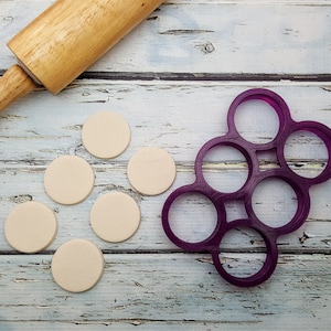 May include: Six round pieces of white clay dough are arranged on a white wooden surface. A purple plastic circle cutter with six circles is to the right of the dough. A wooden rolling pin is to the left of the dough.