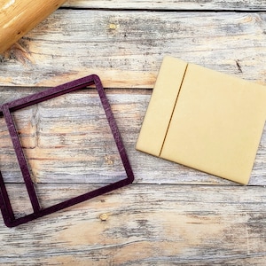 May include: A purple plastic cookie cutter shaped like a picture frame with a smaller rectangle inside. A square piece of dough is next to the cutter on a wooden surface.
