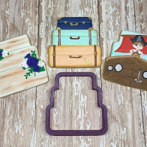 May include: An assortment of decorated sugar cookies in the shapes of a wedding cake, stacked suitcases, a pirate ship, and a cookie cutter. The cookies are decorated with various colors, including blue, white, and brown. The suitcases have a tag that says "Barton".
