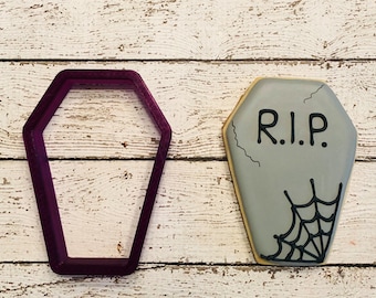 May include: A purple plastic cookie cutter shaped like a coffin. Next to it is a gray sugar cookie decorated with black icing to look like a tombstone with the words "R.I.P." and a spider web.