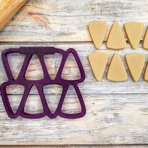 May include: Purple plastic candy corn cookie cutter with the text "Candy Corn 24" on the top. Nine brown candy corn shapes are arranged in two rows on a wooden surface.