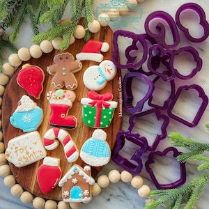 May include: A set of purple Christmas cookie cutters on a wooden board with a variety of decorated Christmas cookies. The cookies include a gingerbread man, a snowman, a present, a stocking, a candy cane, a hot chocolate mug, a light bulb, a gingerbread house, and a tag that says "For Santa".
