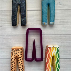 May include: Five decorated sugar cookies shaped like trousers. The cookies are decorated with different patterns, including glitter, denim, leopard print, and stripes. A purple plastic cookie cutter shaped like trousers is also included.