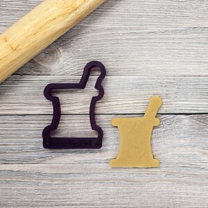 May include: A purple cookie cutter in the shape of a mortar and pestle next to a cookie cut in the same shape. A wooden rolling pin rests in the upper left corner. The objects are on a light wood-grain surface.