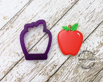 Peut inclure: Emporte-pièce en plastique violet en forme de pomme avec le texte "Tall Apples" en bas. À côté de l'emporte-pièce se trouve un biscuit en forme de pomme rouge avec une tige et des feuilles vertes.