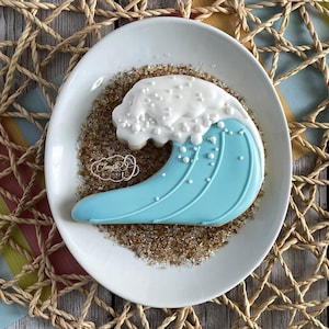 May include: A blue and white sugar cookie decorated to look like a wave crashing on a beach. The cookie is on a white plate with brown sugar sprinkles.