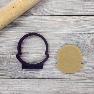 May include: A purple plastic cookie cutter shaped like a shield with a circular top and the text "Squadron 35". A round, tan cookie is next to the cutter. A wooden rolling pin is in the background.