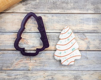 May include: A purple plastic Christmas tree cookie cutter with the words "Snack Tree" on the side, next to a white iced Christmas tree cookie with red and green sprinkles.