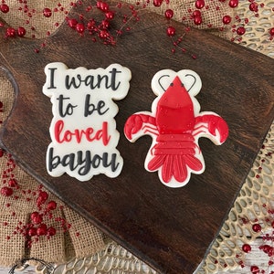 K&ouml;nnte beinhalten: Zwei verzierte Zuckerkekse auf einem Holzbrett. Ein Keks ist rot und hat die Form eines Krebses. Der andere Keks ist wei&szlig; mit schwarzem Zuckerguss und dem Text "I want to be loved bayou".