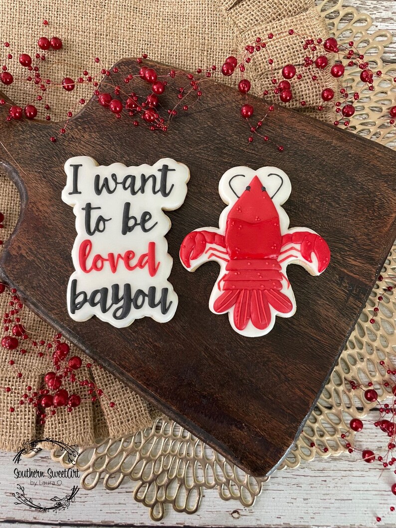 K&ouml;nnte beinhalten: Zwei verzierte Zuckerkekse auf einem Holzbrett. Ein Keks ist rot und hat die Form eines Krebses. Der andere Keks ist wei&szlig; mit schwarzem Zuckerguss und dem Text "I want to be loved bayou".