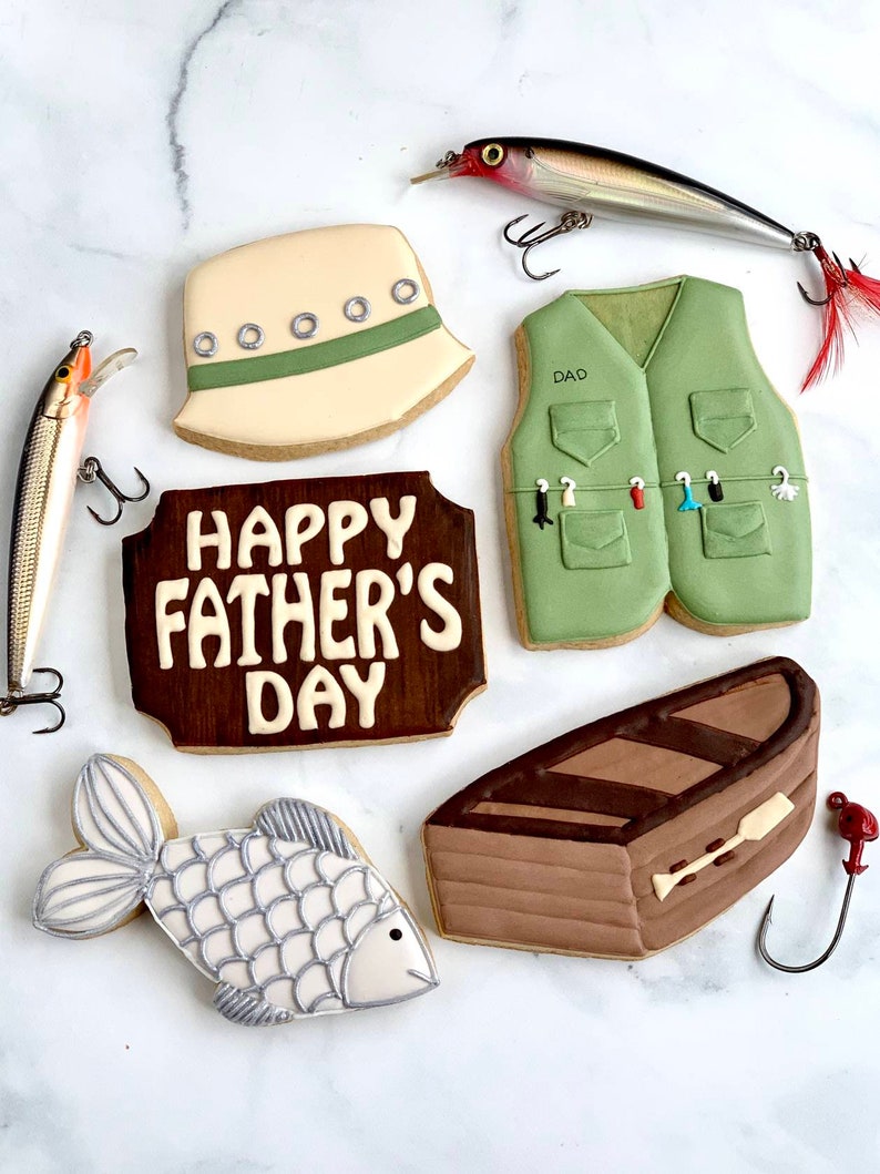 May include: An assortment of decorated sugar cookies with a fishing theme. The cookies include a fishing vest, a boat, a fish, a fishing hat, and a sign that says "Happy Father's Day". Two fishing lures are also present.