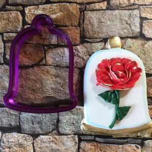May include: Purple plastic cookie cutter shaped like a glass dome with a red rose inside. The cookie cutter is next to a decorated cookie with a red rose inside a glass dome.