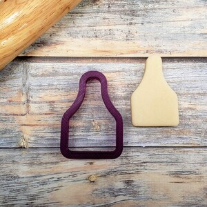 May include: A purple plastic cookie cutter shaped like a cattle ear tag, next to a light brown cookie cut out in the same shape on a wooden surface.