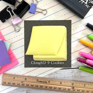 May include: A yellow sugar cookie decorated to look like a sticky note. The cookie is on a black background with the text "Clough'D 9 Cookies".