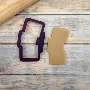 May include: Purple plastic cookie cutter shaped like a small rectangular box with a rounded top. A brown cookie is cut out using the cookie cutter.
